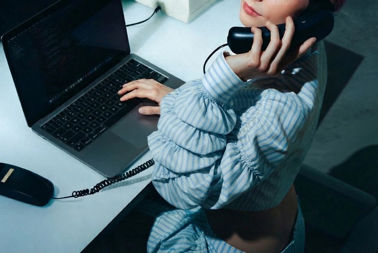 An IT help desk worker at a laptop takes a call on a corded desk phone, her face out of frame